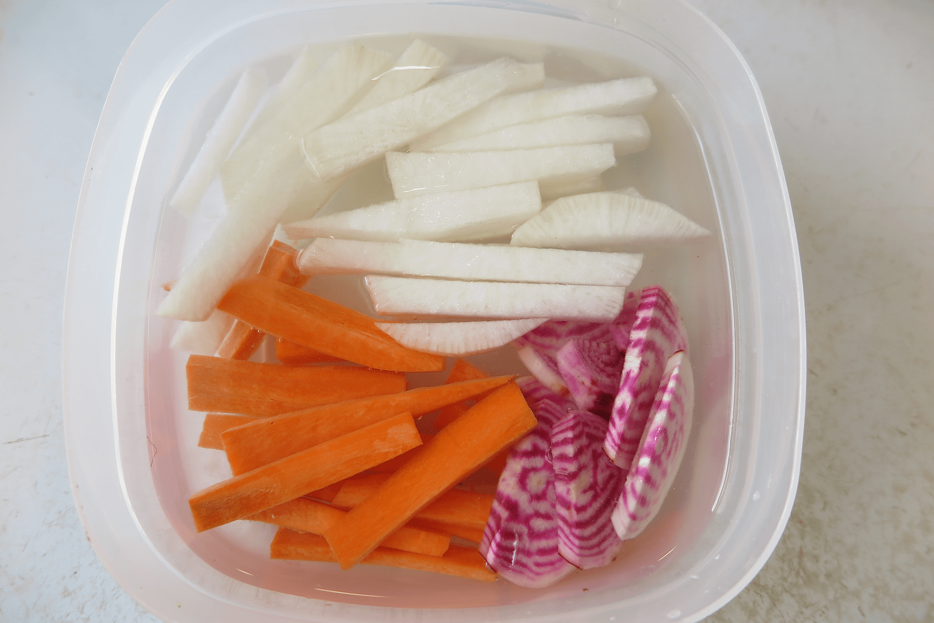 Sliced root vegetables submerged in water in a tupperware container