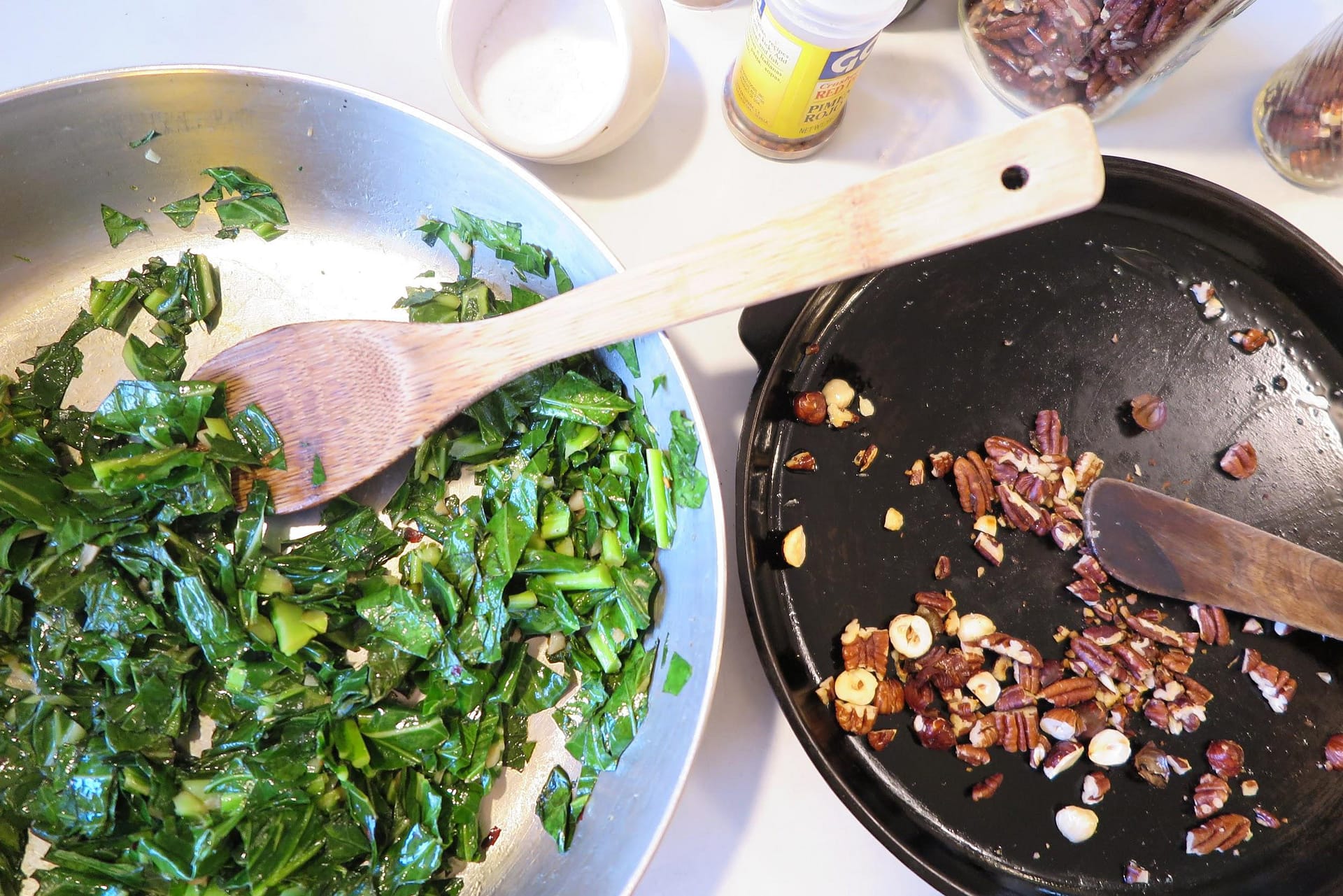 Silver sautee pan with leafy greens and a wooden spoon on the left. On the right, a cast iron pan with toasted chopped nuts.