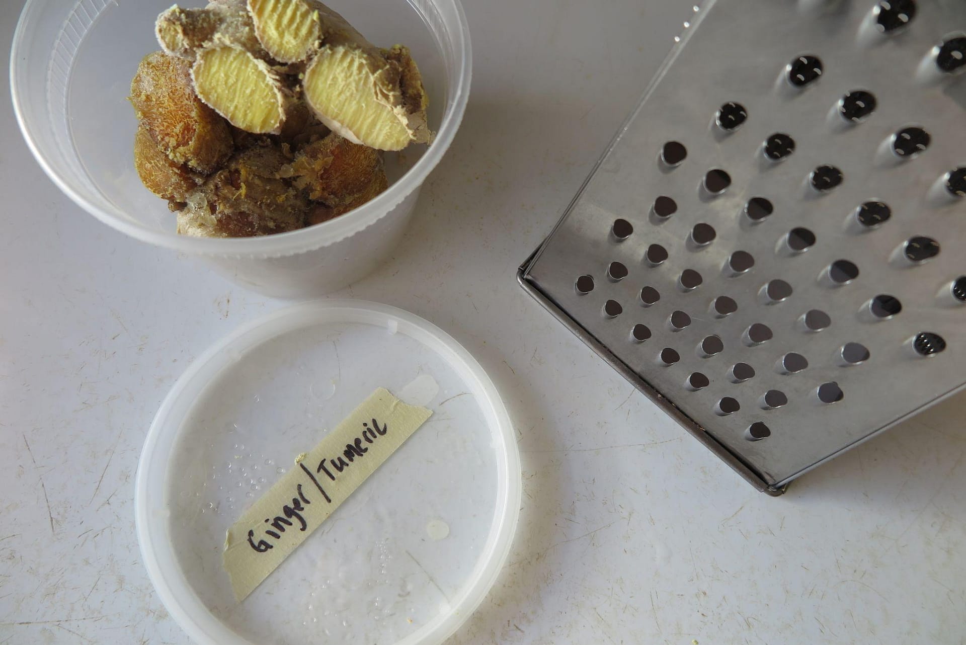 A plastic container of ginger root sitting on a white counter next to a box grater