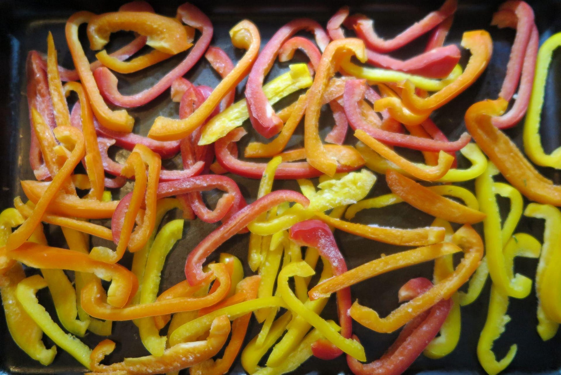 A dark cookie sheet of slices of orange, yellow, and red bell peppers