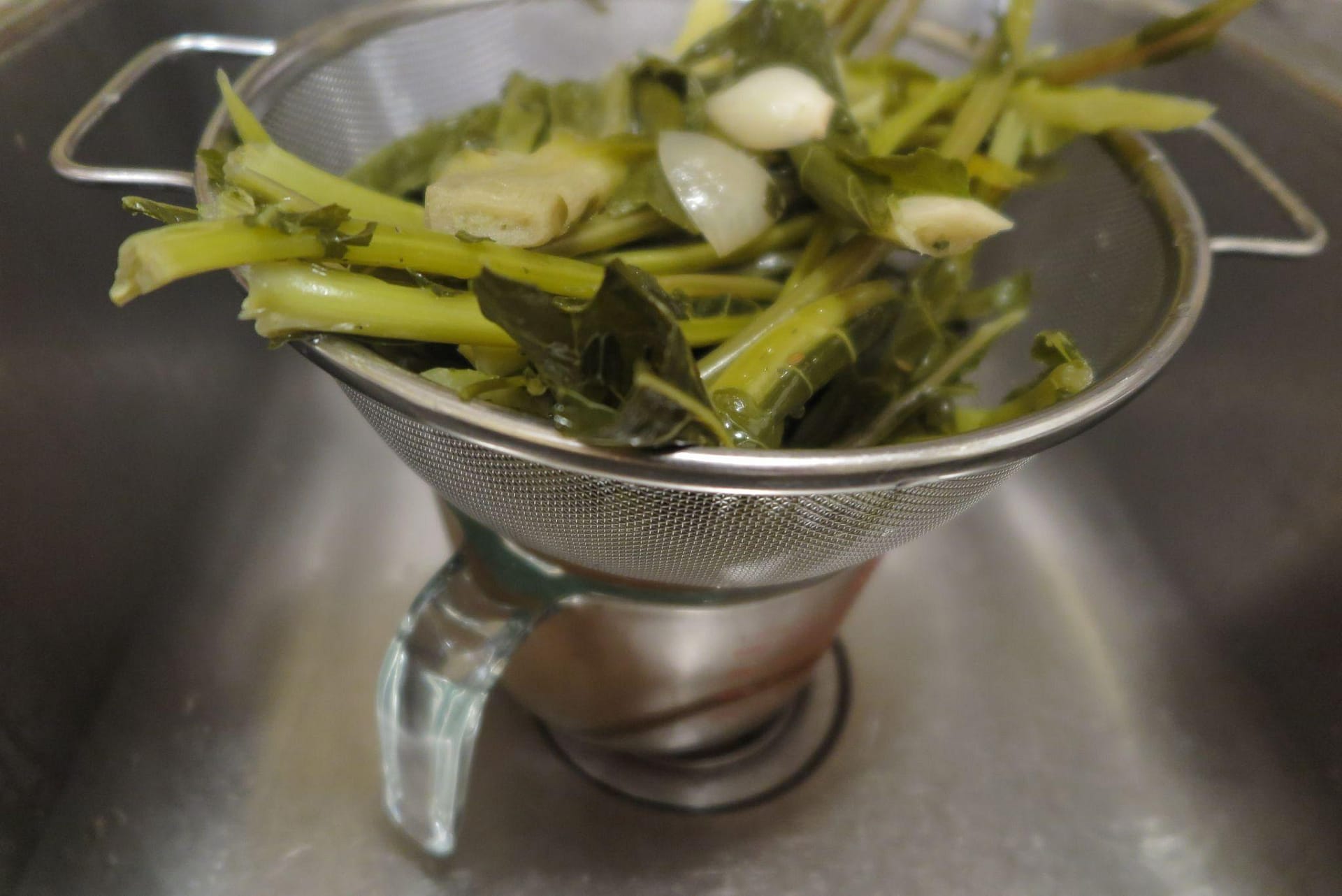 Metal strainer of cooked veggie scraps set on a large glass measuring cup in a sink