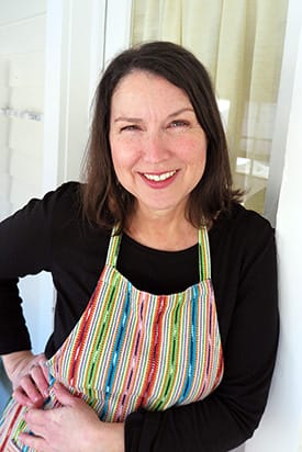 Photo of brunette woman in colorful striped apron leaning against white wall