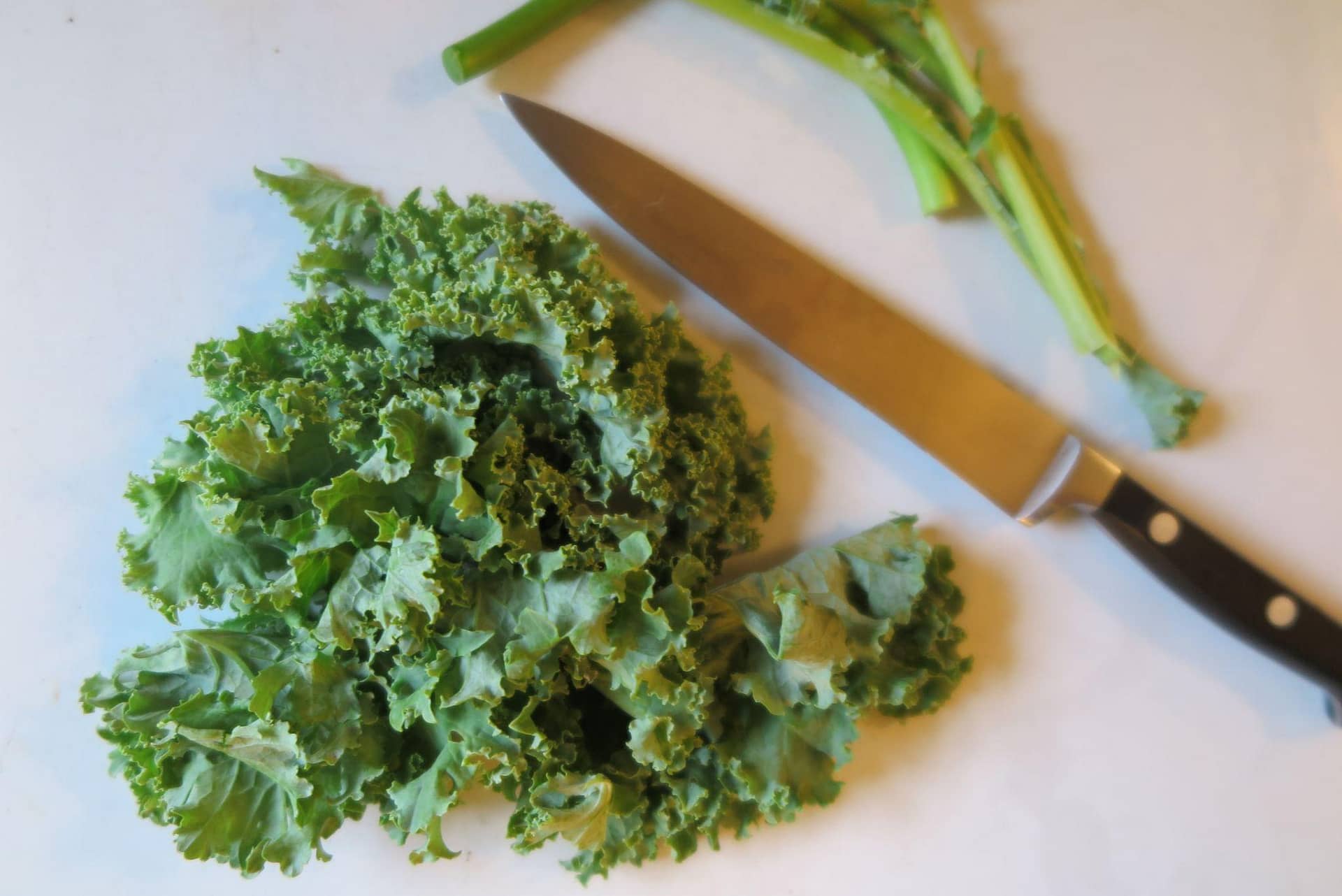 Chopped kale on a white background next to a kitchen knife and garlic scapes