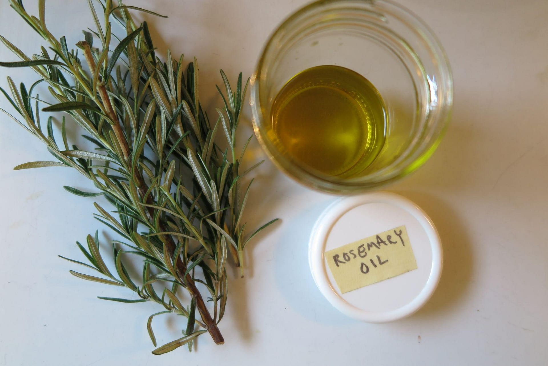 A bundle of rosemary next to a small glass jar of yellow oil with the lid removed labeled rosemary oil