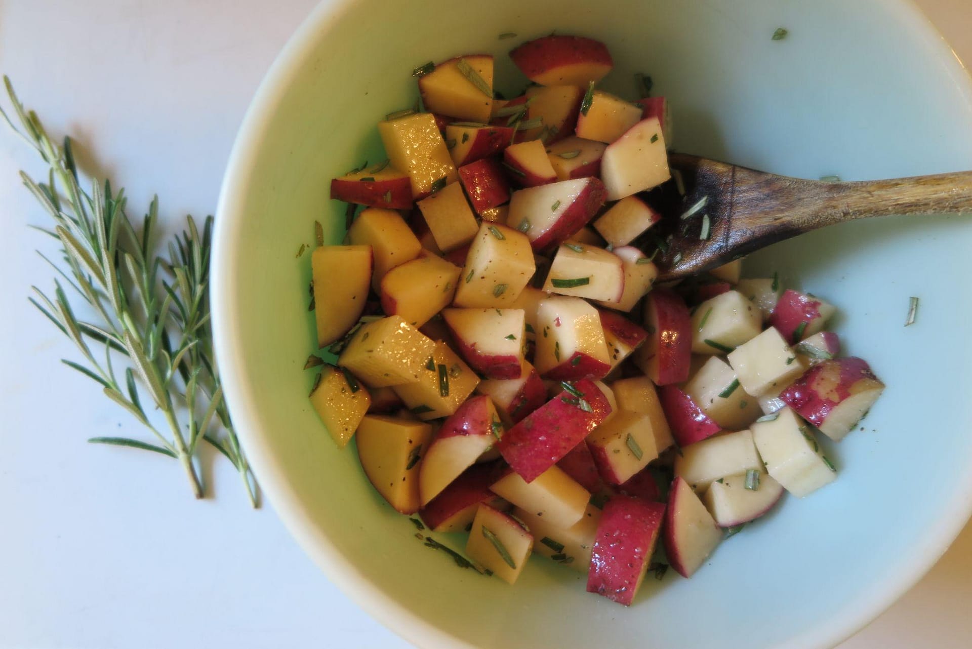 Cut uncooked potatoes tossed in oil and rosemary in a pale green bowl with a wooden spoon