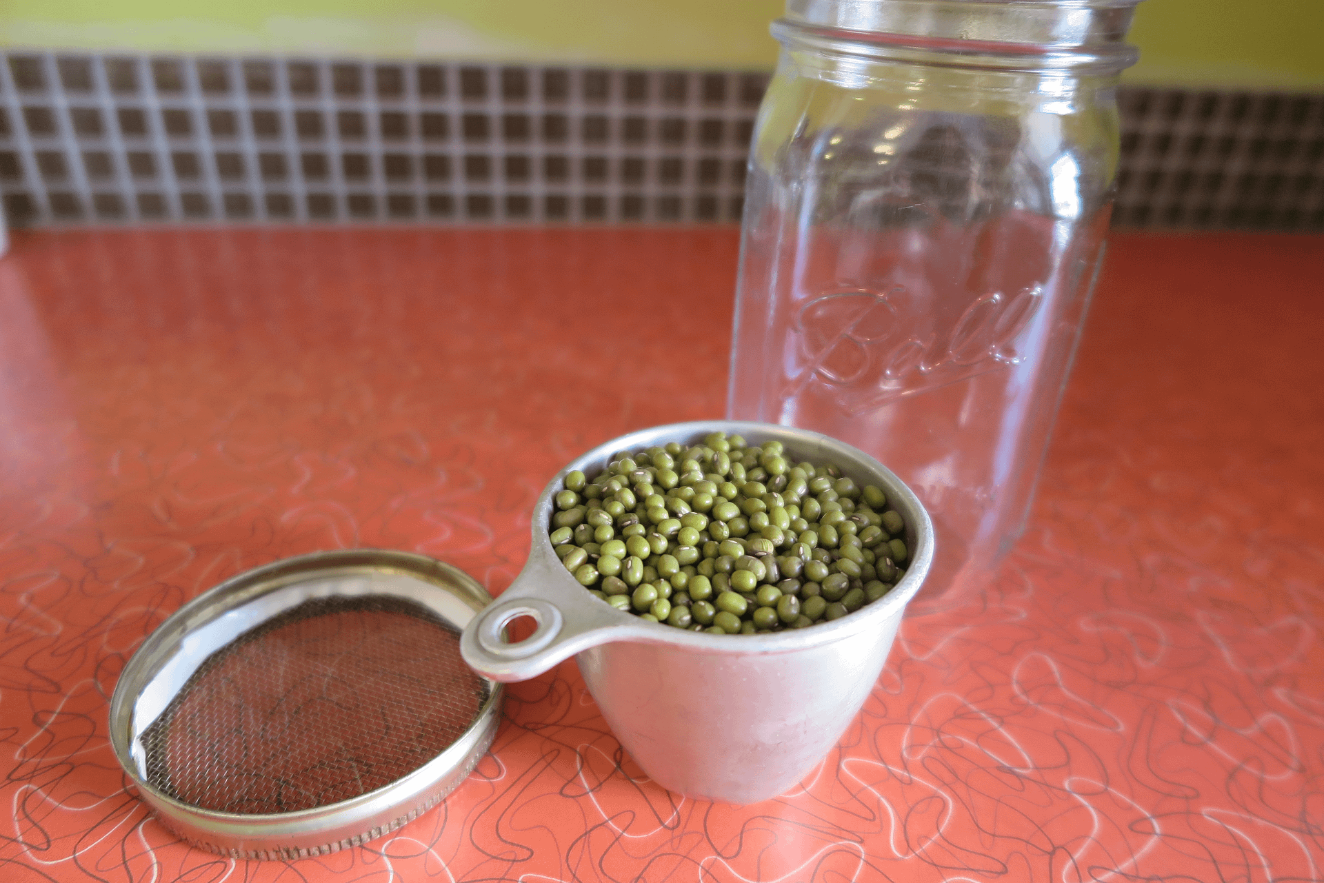 An empty mason jar, a metal measuring cup, and a mason jar lid ring with a mesh screen taped in sitting on a red counter