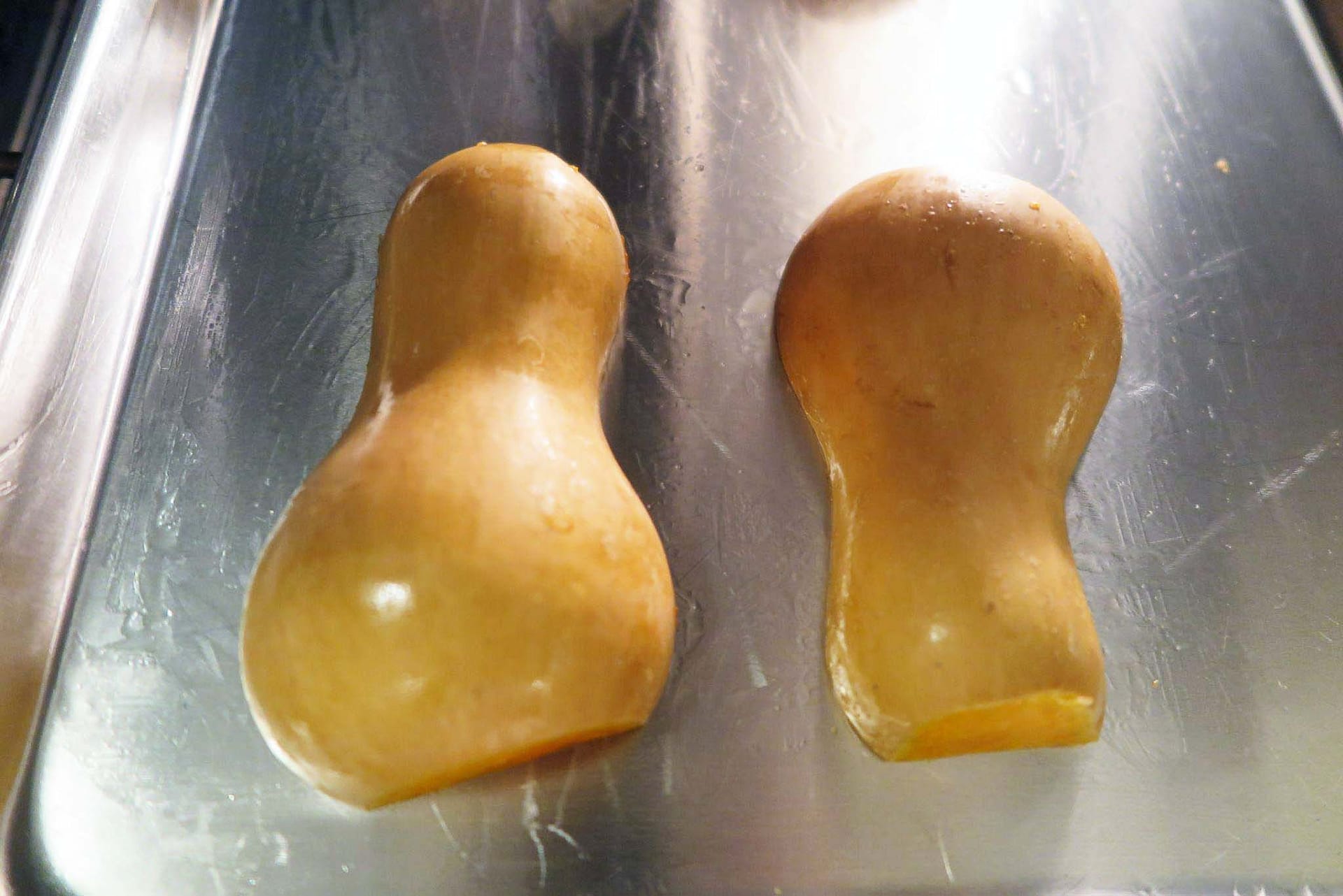 A butternut squash, on a silver baking sheet, sliced in half skin side up, uncooked